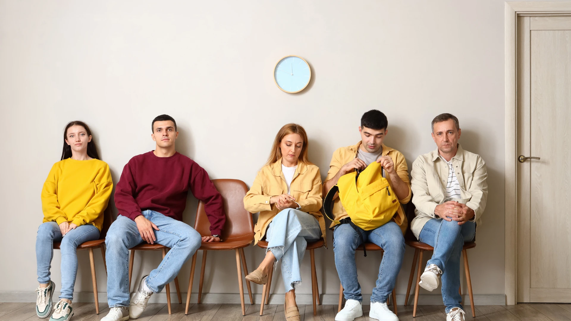 A group of five people seated on chairs, facing a plain wall in a casual setting.