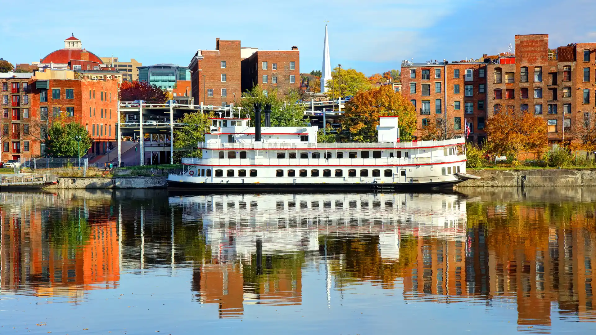 Historic riverfront in Troy, New York, featuring a white riverboat docked along the waterfront with brick buildings, autumn trees, and reflections on the calm Hudson River.