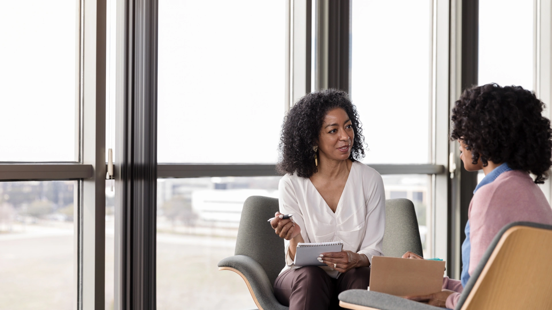 Two women engaged in conversation while seated in chairs, sharing smiles and attentive expressions.